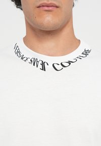 Balta medvilninė marškinėliai su juodu spausdintu grafiniu elementu, kuris aplink apykaklę yra užrašytas kreivėmis „VERSACE JEANS COUTURE“.}}