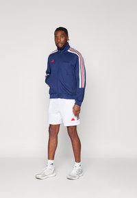 Giacca da track zip-up blu navy con accenti rossi e strisce bianche; abbinata a pantaloncini bianchi e scarpe da ginnastica grigie. Calzini bianchi sono visibili.