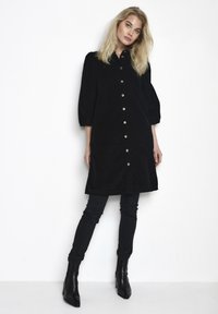 Abito in corduroy nero con bottoni, colletto e maniche a sbuffo, con dettagli in bottoni argentati. Abbinato a pantaloni neri attillati e stivaletti.