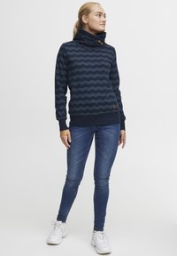 Mørkeblå genser med høy krage, med bølgemønster og treknapper. Kombinert med blå jeans og hvite joggesko.