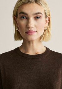 Ruskea neulottu pusero pyöreällä pääntiellä, jossa sileä pinta. Malli käyttää kultaisia renkkikorvakoruja ja hänellä on suora polkkatukka.