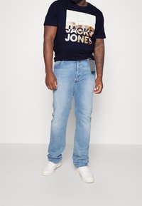 Granatowa koszulka z logo "Jack & Jones" w kolorze złotym, zestawiona z jasnoniebieskimi dżinsami. Białe sneakersy o gładkiej fakturze. Prezentuje swobodny wygląd.