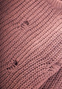 Pletený svetr v jemně růžovém odstínu s texturovaným vzorem a několika malými otvory, které naznačují opotřebení nebo design ve stylu distressed.