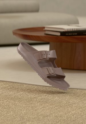 Hellbrauner Slip-On-Sandal mit zwei verstellbaren Schnallenriemen, liegend auf einem gewebten beigen Teppich in der Nähe eines hölzernen Couchtisches mit Büchern.