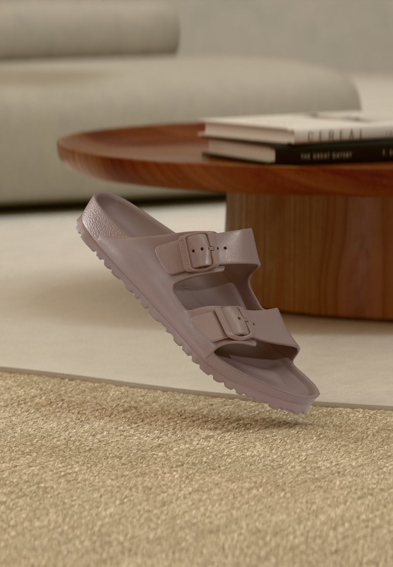 Ανοιχτό καφέ σανδάλι slip-on με δύο ρυθμιζόμενες πόρπες, τοποθετημένο σε υφαντό μπεζ χαλί κοντά σε ξύλινο τραπεζάκι σαλονιού με βιβλία.