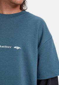 Camiseta de manga corta de algodón en color verde azulado, con el logo en blanco de "Quiksilver" y un gráfico estilizado. Textura suave con un diseño de corte relajado.