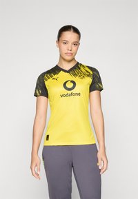 Maillot de football jaune avec des accents noirs, présentant un col en V et un design graphique sur les manches. Logo et sponsor imprimés à l'avant.
