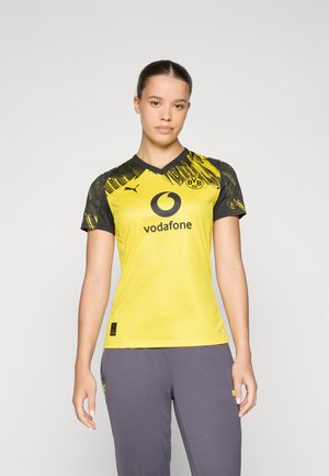 Gul fodboldtrøje med sorte detaljer, med V-hals og et grafisk design på ærmerne. Logo og sponsor trykt på fronten.