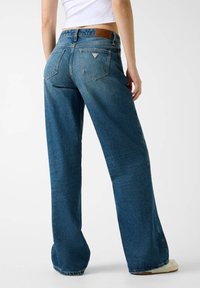 Niebieskie dżinsy o szerokich nogawkach, z wysokim stanem, pięcioma kieszeniami oraz naszywką z logo na tylnej części pasa. Tkanina wydaje się solidna, o teksturowanym wykończeniu.