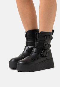 Bottines noires en similicuir avec trois sangles réglables à boucle, fermetures éclair latérales et semelles compensées épaisses. Texture lisse et design durable.