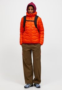 Person in orange gestepptem Jacke, kastanienbraunem Kapuzenpullover, braunen Cargo-Hosen, blau-schwarzen Turnschuhen und schwarzem Rucksack steht vor weißem Hintergrund.