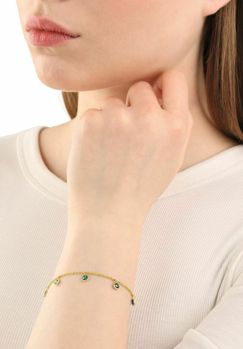 Pulsera de cadena de oro con múltiples pequeñas piedras ovaladas de color turquesa, llevada en una muñeca, sobre una blusa de color claro.