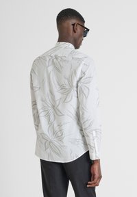 Camisa ligera de manga larga con un patrón floral gris sobre un fondo blanco, con un dobladillo curvado y puños con botones.