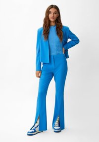 Abito su misura blu brillante composto da un blazer aderente con una superficie liscia, pantaloni svasati con fessure laterali e sneakers blu con accenti bianchi.