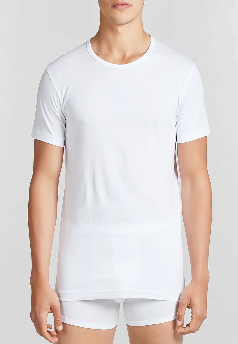T-shirt bianca a maniche corte realizzata in tessuto liscio, con scollatura rotonda e vestibilità regolare. Indossata su pantaloni corti bianchi aderenti.