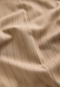 Tessuto beige con una sottile texture, caratterizzato da sottili righe verticali nere. Il materiale ha un aspetto liscio e tessuto.