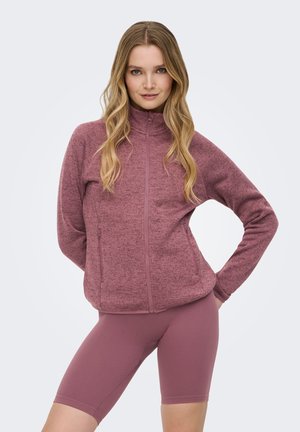 Pitkähiuksinen nainen, jolla on yllään vetoketjulla suljettava vaaleanvioletti fleecetakki ja sitä vastaavat ribbineulotut pyöräilyshortsit, seisoo käsi lanteilla.