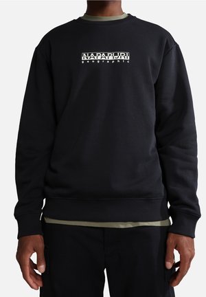 Person iført sort Napapijri sweatshirt med hvidt logotekst centreret på brystet, lagdelt over olivengrøn skjorte.