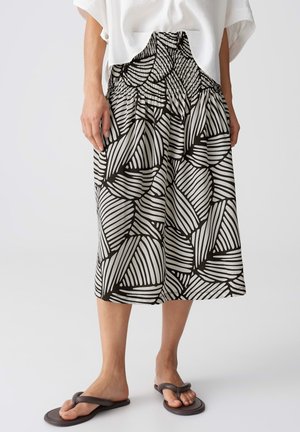 Vrouw die een zwart-witte midi rok met bladdessin draagt, een wit blouse en donkerbruine teenslippers, staande op een effen achtergrond.