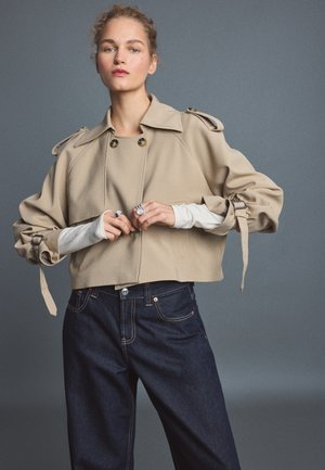 Jeune femme portant une veste courte beige avec des lanières à boucle et un jean taille haute foncé sur un fond gris uni.