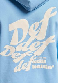 Lyseblå hettegenser med store hvite teksttrykk, inkludert "Def," "def," og "still ballin' since 2006." Myk stoff med glatt tekstur.