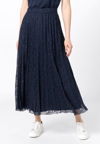 Námořnicky modrá plisovaná maxi sukně z krajky s texturovaným vzorem, se střiženým pasem a průhledným lemem, doplněná bílými teniskami.