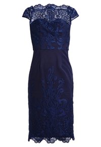 Tummansininen mekko, jossa on pitsinen yläosa kukka- brodeerauksella, lyhyet hihat ja vartalonmyötäinen siluetti, joka päättyy aaltoilevaan helmaan.