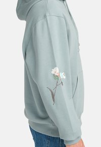 Light blue hoodie made from soft material. Features an embroidered floral design on the left sleeve. Kangaroo pocket in front.