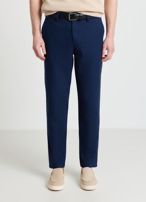 Homme portant un pantalon slim bleu marine, une ceinture en cuir noire, un haut en maille beige et des chaussures beiges à enfiler, debout devant un fond uni.