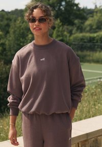 Smeđa majica s rebrastim manšetama i rubom, s bijelim natpisom "self."; uparena sa pripadajućim trenirkama. Mekani, ležerni materijal, vanjski prostor.