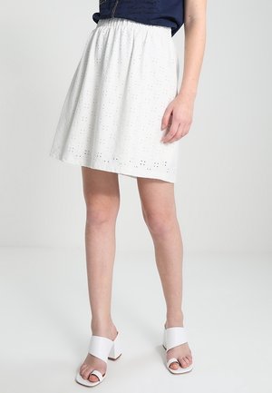 Femme portant une jupe blanche à œillets, un haut bleu marine et des sandales blanches à talons blocs ouverts, debout sur un fond clair uni.