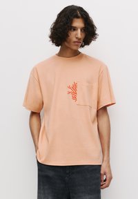 Tricou din bumbac piersică cu mâneci scurte, având un buzunar și un design abstract portocalie pe piept. Textură netedă, croială lejeră.