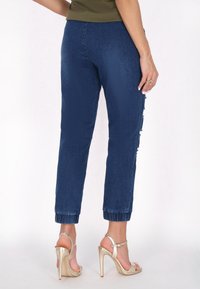 Sötétkék farmer tapered nadrág, szakadozott oldalsó részletekkel és felhajtott szegéllyel, világos arany pántos magassarkú szandálokkal párosítva.