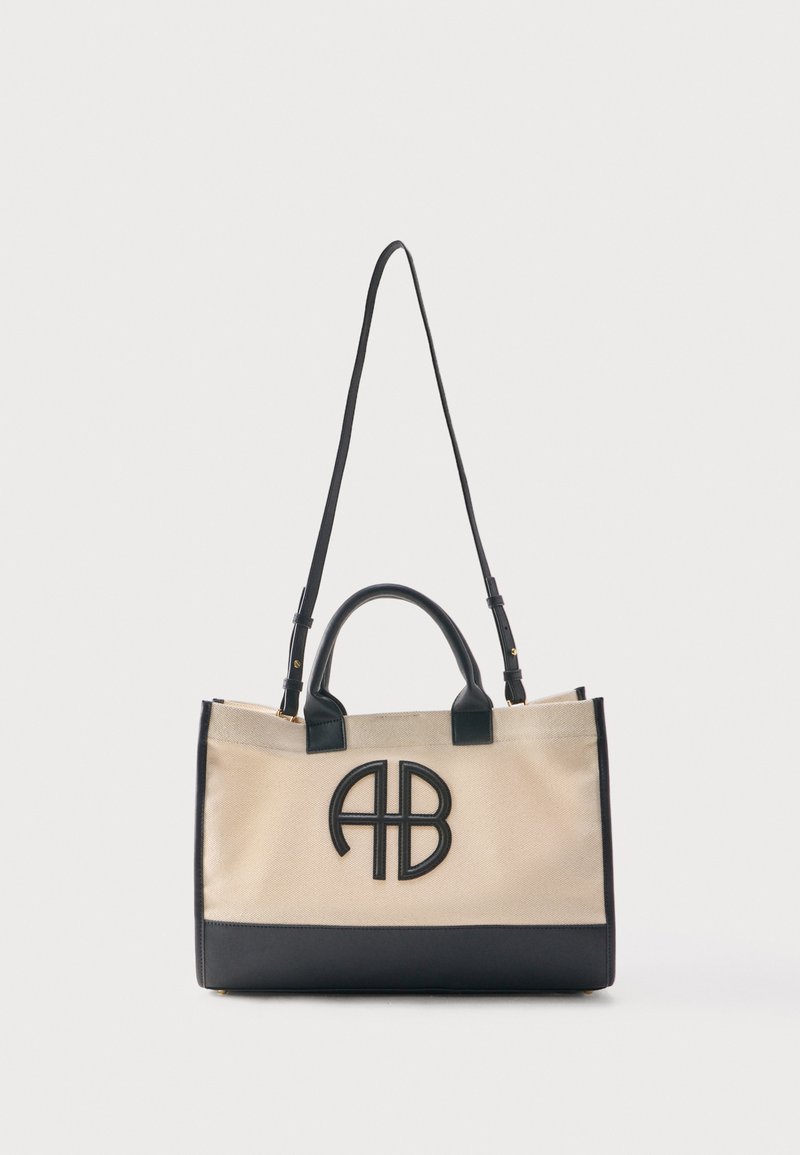 Μπεζ καμβά τσάντα ώμου με μαύρες δερμάτινες λαβές, διακοσμητικά και ιμάντα ώμου, με μαύρο λογότυπο "AB" στο μπροστινό μέρος.