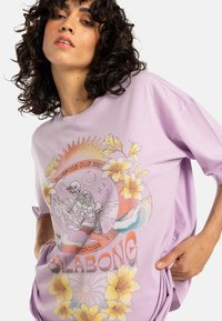 Tricou din bumbac violet cu o croială supradimensionată, având un grafic colorat cu un schelet care face surf printre valuri și flori, cu textul "Surfing Our Way."