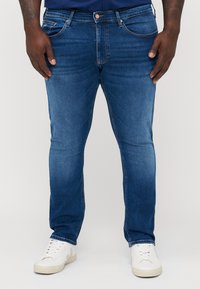 Blå denimjeans med smal passform, fem fickor, kopparfärgade detaljer och subtil blekning. Matchas med vita sneakers för kontrast.