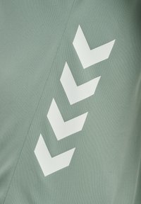 Grön tygblus med en texturerad yta och ett stort vitt chevronmönster på ena sidan, vilket skapar en kontrast i designen.