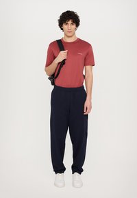 Röd t-shirt med kort ärm och logotyp, kombinerad med marinblå joggingsbyxor med sidofickor. Skor: vita sneakers. Modellen står rak med en väska.