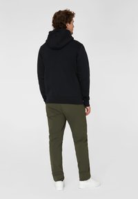 Svart huva sweatshirt med texturerad tyg, matchad med olivgröna byxor. Personen har på sig vita sneakers utan synliga logotyper.