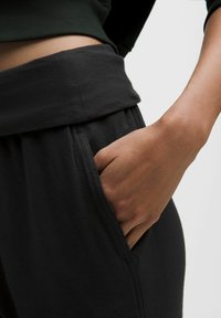 Fekete sportnadrág széles rugalmas derékpánttal, oldalzsebbel és puha, texturált anyagból. A modell keze laza mozdulattal pihen a zsebben.