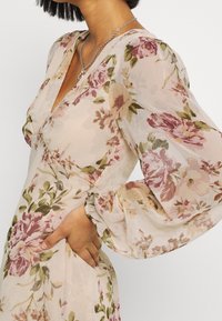 Vestido floral de tela transparente, con patrones en rosa y verde. Diseño en forma de V y mangas amplias con detalles en los puños. Textura suave.