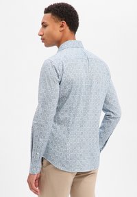 Niebieska koszula z długim rękawem i klasycznym kołnierzykiem, z delikatnym geometrycznym wzorem i mankietami na guziki. Noszona z jasnymi spodniami.