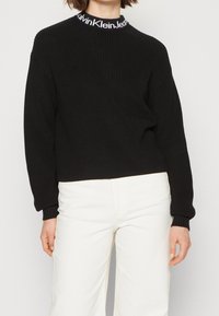 Czarny sweter dziergany o prążkowanej fakturze z okrągłym dekoltem, z białym logo marki na kołnierzu. W zestawie z białymi spodniami.