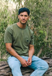 Green cotton T-shirt with a chest pocket featuring an elephant design. Paired with light blue jeans. Natural outdoor setting in the background.