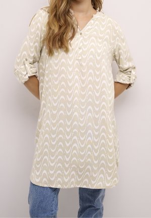 Femme portant une tunique beige à manches longues avec des motifs ondulés blancs et un jean bleu, manches retroussées et mains dans le dos.