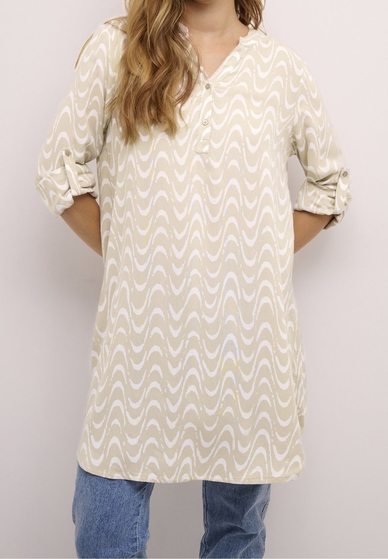 Femme portant une tunique beige à manches longues avec des motifs ondulés blancs et un jean bleu, manches retroussées et mains dans le dos.