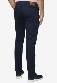 Muž oblečený v tmavě modrých slim-fit kalhotách a modrých teniskách, stojící proti bílému pozadí, zobrazený zezadu.