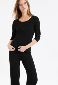 Kobieta w czarnym prążkowanym topie z długim rękawem i dopasowanych spodniach, poprawiająca dół ubrania, stojąca na tle gładkiej białej ściany.