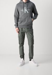 Szara bluza z kapturem z białym logo, zestawiona z zielonymi spodniami typu cargo z bocznymi kieszeniami oraz białymi trampkami. Prosty design i luźne dopasowanie.