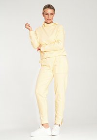 Żółta bluza z kapturem i pasujące spodnie wykonane z miękkiego materiału, z wysokim kołnierzem i bocznymi kieszeniami, w połączeniu z białymi trampkami i przetarciem po bokach.